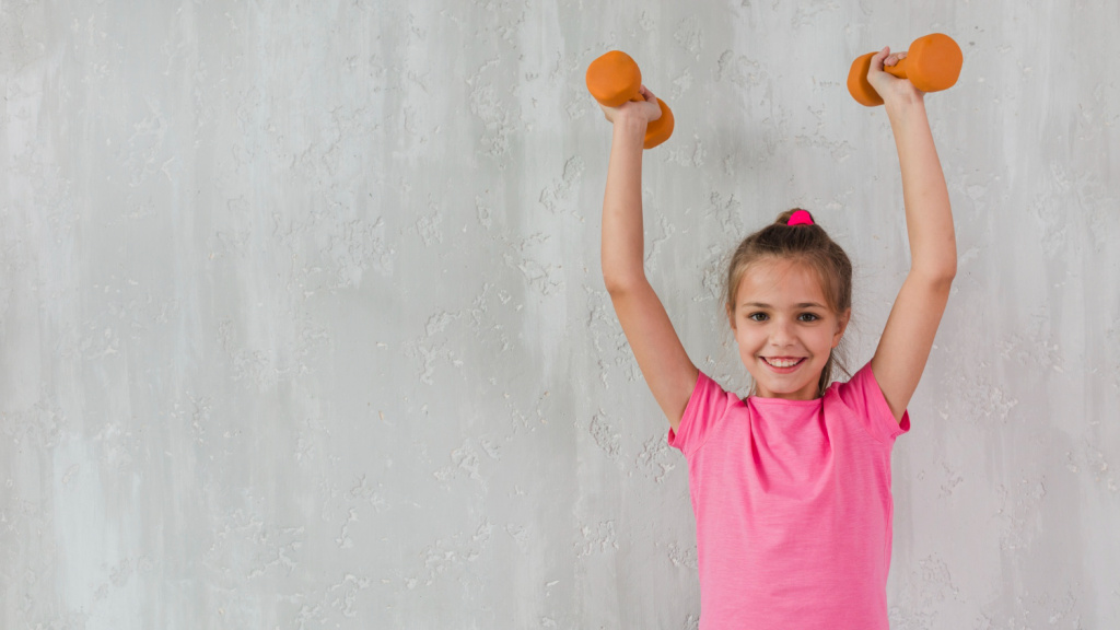portrait-of-a-smiling-girl-raising-her-hands-holding-dumbbells-in-front-of-concrete-wall.jpg